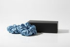 Two silk scrunchies and a closed matte black rectangular box arranged on a clean white surface. The scrunchies are sky blue, voluminous, and softly gathered to show the smooth, glossy silk texture. The black box is minimal with no text or logo. Soft, even studio lighting creates gentle shadows and highlights the sheen of the silk. Clean, modern, and elegant composition.
